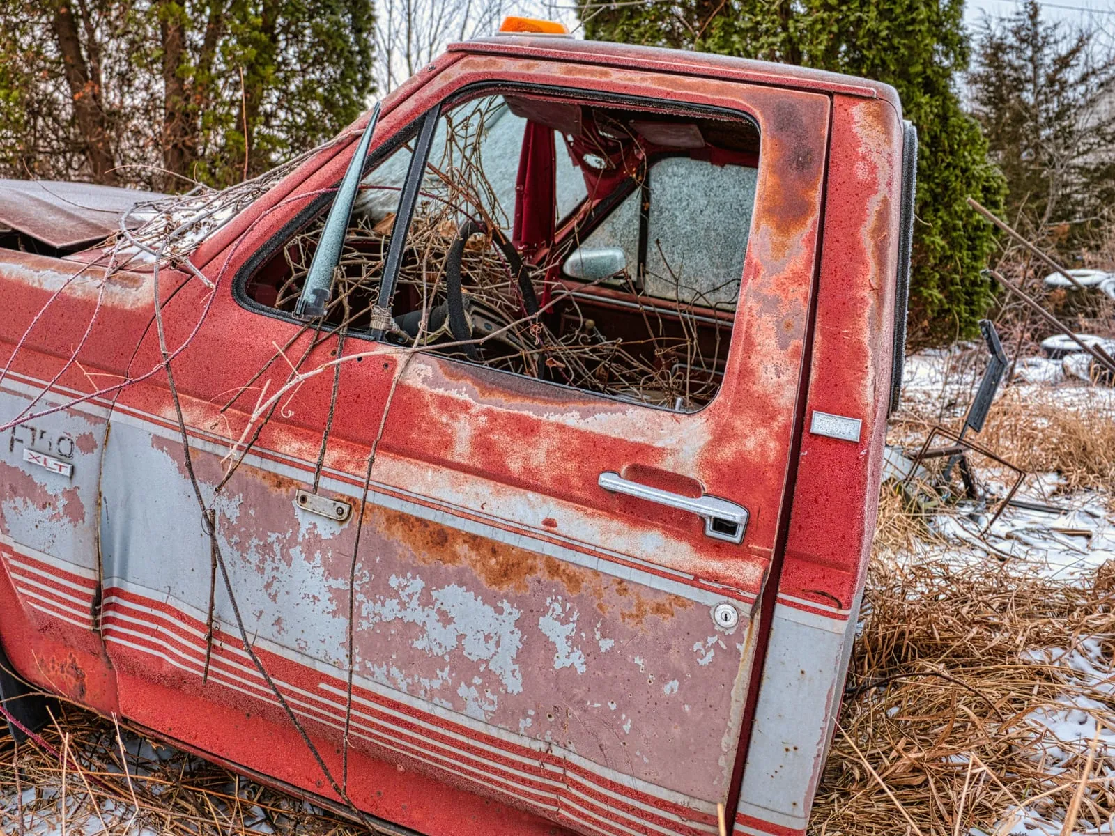 Camioneta pickup roja y oxidada con una ventana rota, estacionada sobre pasto seco con un poco de nieve